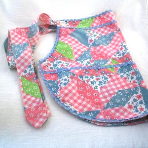 May include: A colorful patchwork apron with a matching tie. The apron features a mix of pink gingham, floral, and solid blue fabrics. The edges are trimmed with a light blue scalloped trim. The tie is made of the same patchwork fabric.