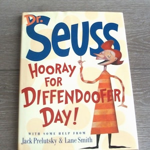 May include: A hardcover book titled "Dr. Seuss Hooray for Diffendoofer Day!" features a cartoon illustration of a character pointing upwards. The title is in large blue and red letters. The book's cover is cream-colored.