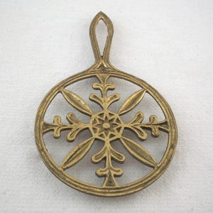 May include: A gold-colored trivet with a handle. The trivet features a circular design with an intricate floral pattern in the center and around the edges. The handle is arched, allowing for easy hanging or storage. The trivet is made of metal.