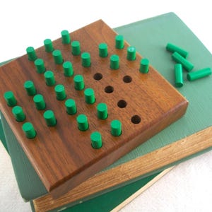 May include: A wooden peg game with a dark brown base and green pegs. The game board is square with holes for the pegs. Several loose green pegs are scattered nearby. The game rests on top of two green books.
