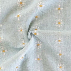 May include: A light blue fabric featuring vertical stripes of embroidered white and yellow daisy-like flowers. The textile has a textured appearance with a soft drape. The word FABLEISM is printed in small white letters near the bottom center.