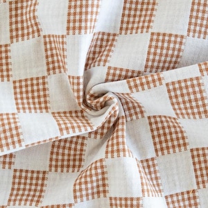 May include: A folded, off-white fabric with a brown checkered pattern. The fabric is soft and has a slight texture, with the checkered design creating a visual grid. The fabric is suitable for home decor.