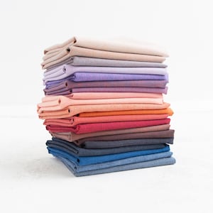 May include: A stack of folded fabric squares in a variety of colors, including beige, purple, pink, orange, red, and blue. The fabric appears to be linen or a similar woven material, with a slightly textured surface. The stack is neatly arranged, showcasing the different hues.