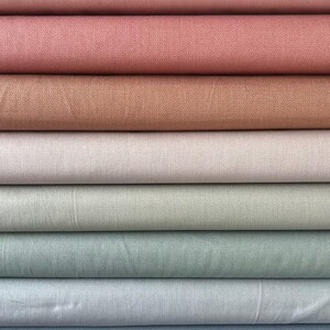 May include: A stack of fabric in various pastel colors, including pink, rose, tan, and blue. Each fabric has a subtle dotted pattern. The fabrics are neatly folded and stacked, showcasing a range of soft, muted tones.