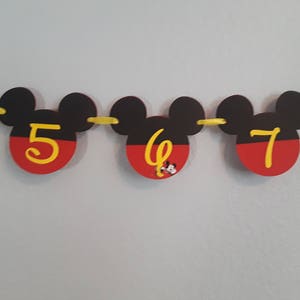 May include: A Mickey Mouse themed birthday banner with black ears and red and yellow circles. The numbers 5, 6, and 7 are written in yellow on the circles.