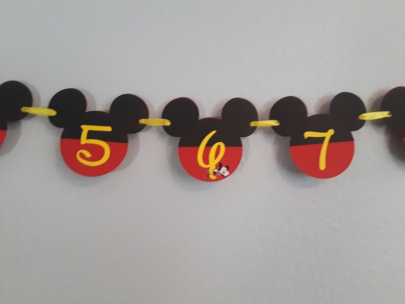 May include: A Mickey Mouse themed birthday banner with black ears and red and yellow circles. The numbers 5, 6, and 7 are written in yellow on the circles.