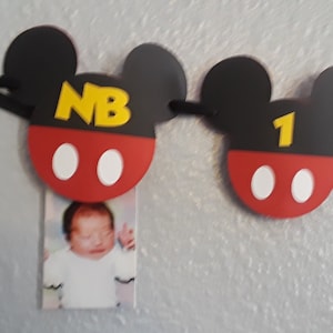 May include: A Mickey Mouse themed banner with black ears, red and white circles, and yellow letters. The banner says "NB" and "1". A photo of a baby is attached to the "NB" section.