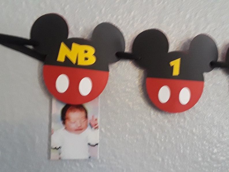 May include: A Mickey Mouse themed banner with black ears, red and white circles, and yellow letters. The banner says "NB" and "1". A photo of a baby is attached to the "NB" section.