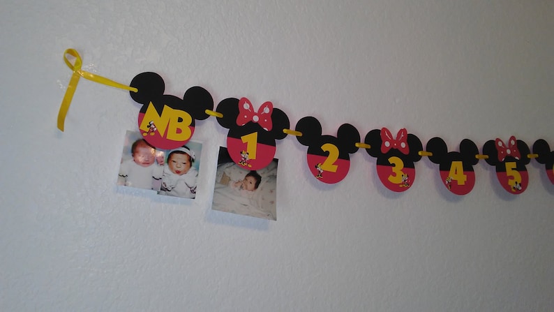 May include: A decorative banner featuring black and red mouse-shaped cutouts strung on a yellow ribbon. The cutouts display yellow numbers from NB to 5, with some featuring red bows and small character graphics. Two square photographs are attached.