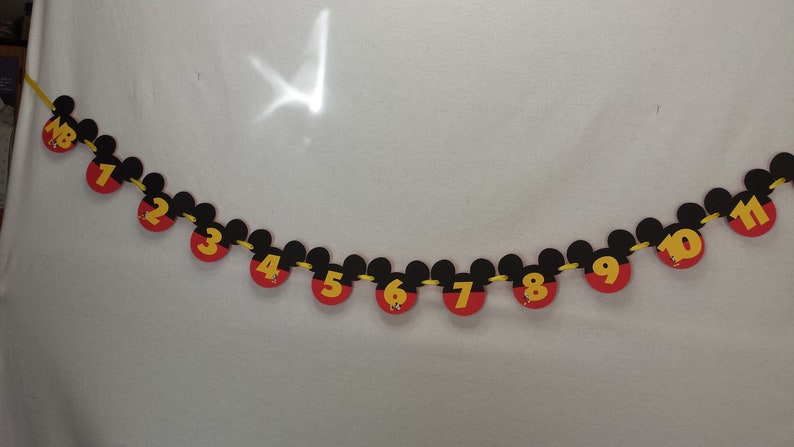 Pu&ograve; includere: Una ghirlanda decorativa composta da una serie di sagome nere a forma di testa di Mickey Mouse infilate su un cordino giallo. Ogni sagoma mostra un numero giallo da 1 a 11, con il primo pezzo che riporta le lettere NB in giallo su sfondo rosso.