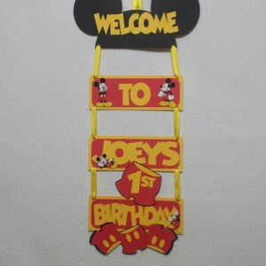 Pu&ograve; includere: Un cartello decorativo sospeso con una sagoma nera delle orecchie di Topolino in alto con il testo giallo WELCOME. Sotto ci sono tre pannelli rettangolari rossi e gialli collegati da nastri gialli che riportano TO, JOEY'S e 1ST BIRTHDAY.