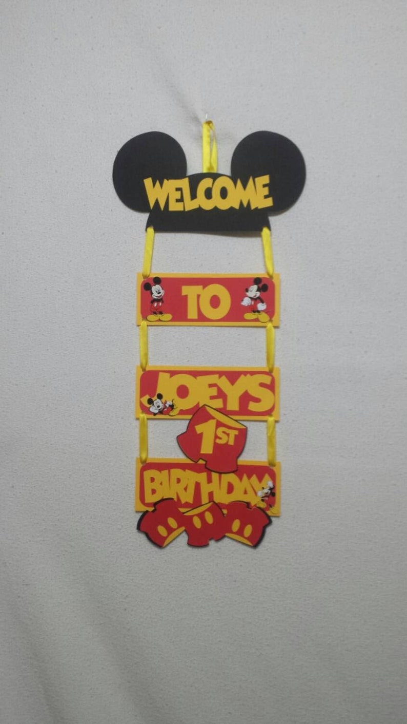 Pu&ograve; includere: Un cartello decorativo sospeso con una sagoma nera delle orecchie di Topolino in alto con il testo giallo WELCOME. Sotto ci sono tre pannelli rettangolari rossi e gialli collegati da nastri gialli che riportano TO, JOEY'S e 1ST BIRTHDAY.