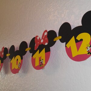 May include: A decorative banner featuring a series of black mouse-ear silhouettes with pink lower sections, strung together on a yellow ribbon. Each cutout displays a large yellow number from six to twelve and a small character illustration.