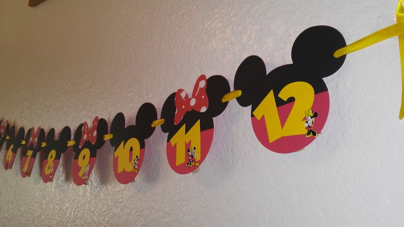 May include: A decorative banner featuring a series of black mouse-ear silhouettes with pink lower sections, strung together on a yellow ribbon. Each cutout displays a large yellow number from six to twelve and a small character illustration.