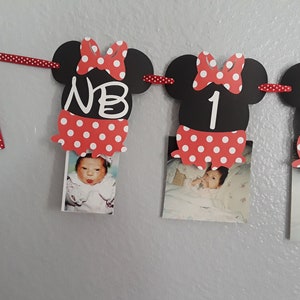 May include: A Minnie Mouse-themed photo banner with black mouse head cutouts, red polka dot bows and skirts, and photos of a baby. The banner includes the letters "NB" and numbers "1" and "2". A red polka dot ribbon is used to hang the banner.