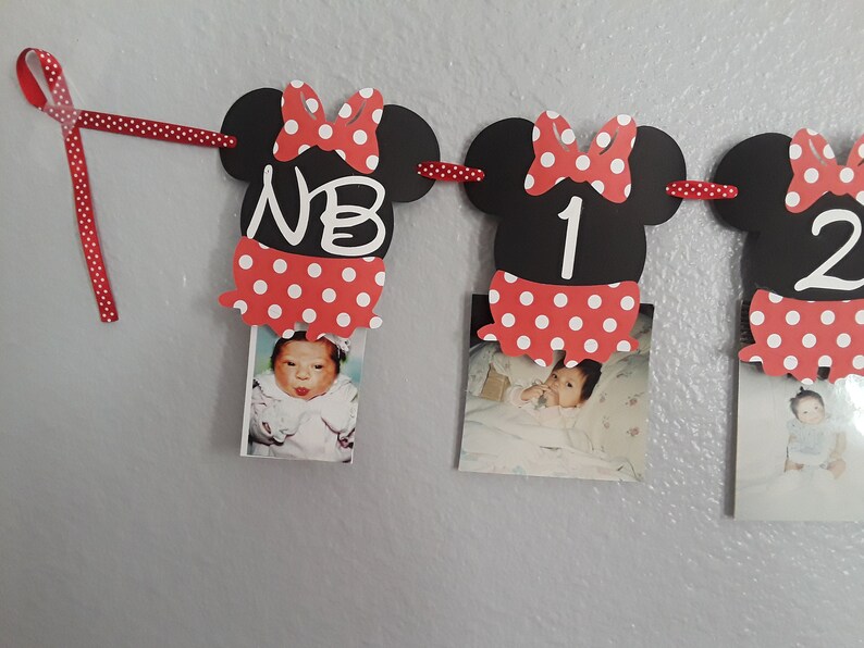 May include: A Minnie Mouse-themed photo banner with black mouse head cutouts, red polka dot bows and skirts, and photos of a baby. The banner includes the letters "NB" and numbers "1" and "2". A red polka dot ribbon is used to hang the banner.