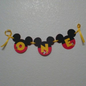 Pu&ograve; includere: Uno striscione decorativo con tre sagome nere a forma di Mickey Mouse collegate da un nastro giallo. Ogni sagoma contiene una grande lettera gialla che forma la parola ONE, con piccoli disegni di personaggi alla base di ogni lettera.