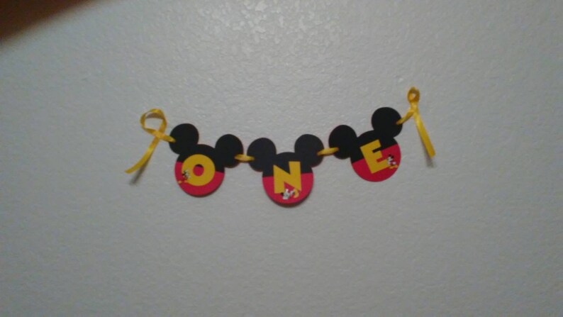 Pu&ograve; includere: Uno striscione decorativo con tre sagome nere a forma di Mickey Mouse collegate da un nastro giallo. Ogni sagoma contiene una grande lettera gialla che forma la parola ONE, con piccoli disegni di personaggi alla base di ogni lettera.