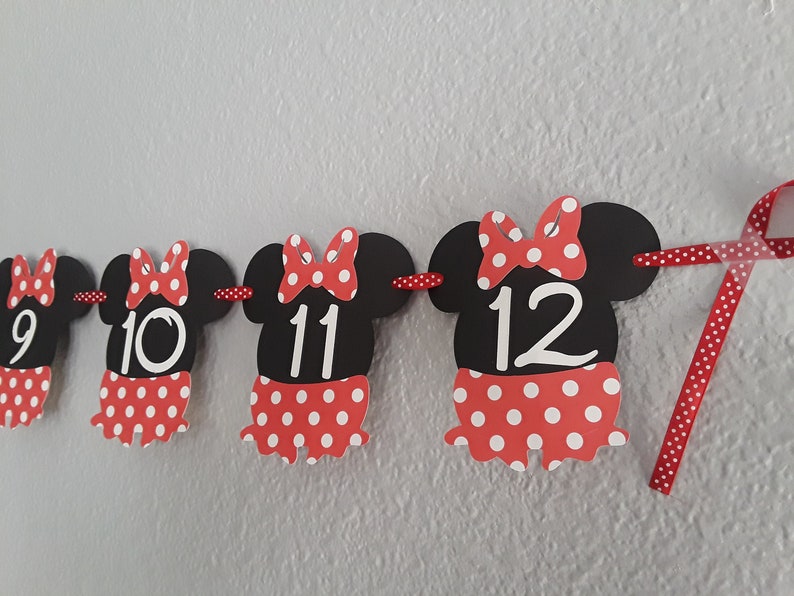 May include: A festive banner featuring black Minnie Mouse-shaped cutouts with red and white polka dot skirts and bows. Numbers 9, 10, 11, and 12 are displayed on the cutouts, perfect for a birthday celebration.