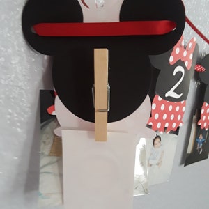 May include: A Minnie Mouse-themed photo banner with black ears, a red ribbon, and a wooden clip. The banner is designed to hold photos, with additional Minnie Mouse cutouts and the number "2" visible.