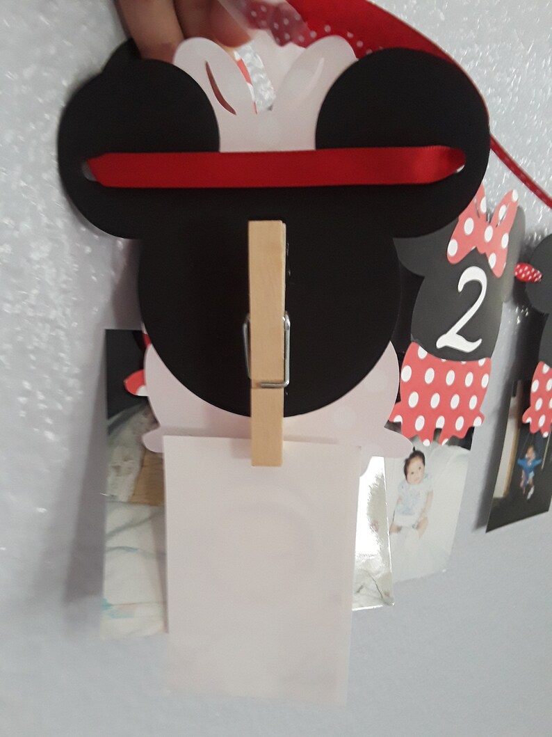 May include: A Minnie Mouse-themed photo banner with black ears, a red ribbon, and a wooden clip. The banner is designed to hold photos, with additional Minnie Mouse cutouts and the number "2" visible.
