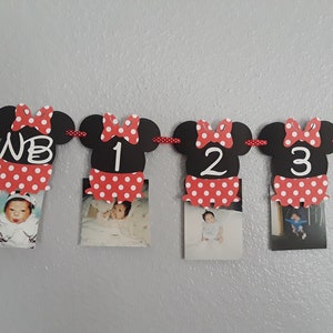 May include: A Minnie Mouse-themed photo banner with black mouse head cutouts, red polka dot bows, and matching skirts. Each cutout holds a baby photo and a number or initials. The banner is strung on a red and white polka dot ribbon.