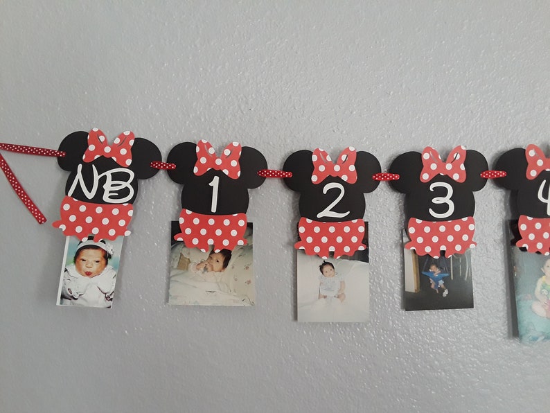May include: A Minnie Mouse-themed photo banner with black mouse head cutouts, red polka dot bows, and matching skirts. Each cutout holds a baby photo and a number or initials. The banner is strung on a red and white polka dot ribbon.