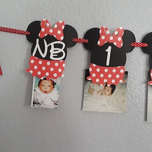 May include: A Minnie Mouse-themed photo banner with black mouse head cutouts, red polka dot bows and skirts, and photos of a baby. The banner includes the letters "NB" and numbers "1" and "2". A red polka dot ribbon is used to hang the banner.