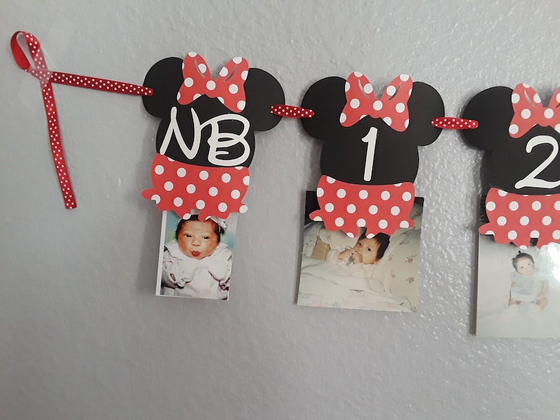 May include: A Minnie Mouse-themed photo banner with black mouse head cutouts, red polka dot bows and skirts, and photos of a baby. The banner includes the letters "NB" and numbers "1" and "2". A red polka dot ribbon is used to hang the banner.