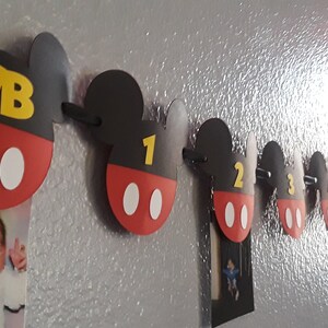 May include: A Mickey Mouse themed baby milestone banner with red, black, and white Mickey Mouse ears and a yellow number on each ear. The banner includes the letters "NB" for newborn, and numbers 1 through 5.