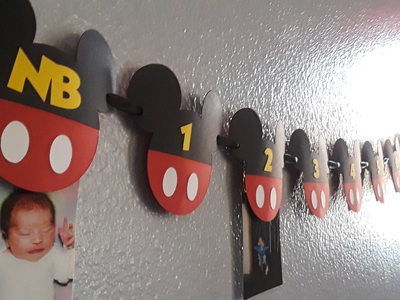 May include: A Mickey Mouse themed baby milestone banner with red, black, and white Mickey Mouse ears and a yellow number on each ear. The banner includes the letters "NB" for newborn, and numbers 1 through 5.