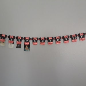 May include: A Minnie Mouse-themed photo banner with red and black elements. Each piece is shaped like Minnie's dress and features a number from NB to 12. Small photos are clipped to each piece, strung together with red string.