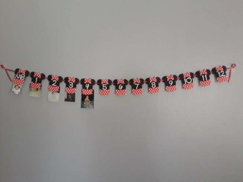 May include: A Minnie Mouse-themed photo banner with red and black elements. Each piece is shaped like Minnie's dress and features a number from NB to 12. Small photos are clipped to each piece, strung together with red string.