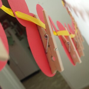 Pu&ograve; includere: Una ghirlanda decorativa con ritagli di carta rossi circolari sospesi a un nastro giallo brillante. Ogni cerchio di carta rossa &egrave; fissato al nastro giallo utilizzando una piccola molletta in legno naturale con una molla in metallo.