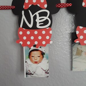 May include: A Minnie Mouse-themed photo banner with black mouse head cutouts, red polka dot bows, and red polka dot skirts. The banner displays the initials "NB" and holds a baby photo. The banner is strung with a red ribbon.