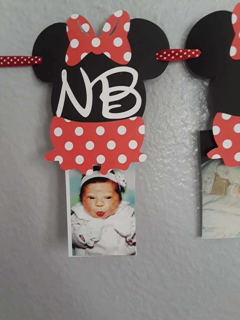 May include: A Minnie Mouse-themed photo banner with black mouse head cutouts, red polka dot bows, and red polka dot skirts. The banner displays the initials "NB" and holds a baby photo. The banner is strung with a red ribbon.