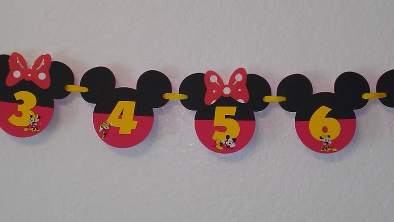 May include: A decorative banner featuring black Mickey Mouse-shaped silhouettes with red bottoms and yellow numbers 3, 4, 5, and 6. Some silhouettes feature a red bow with white polka dots and small illustrations of Minnie Mouse at the base of the numbers.