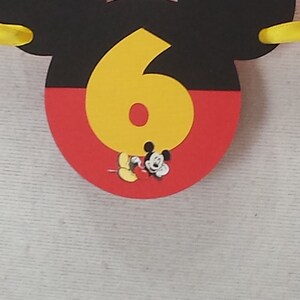 Pu&ograve; includere: Una sagoma decorativa a forma di testa di Topolino, con la parte superiore nera e quella inferiore rossa. Un grande numero sei giallo &egrave; centrato sulla parte anteriore, con una piccola illustrazione di Topolino alla base del numero.
