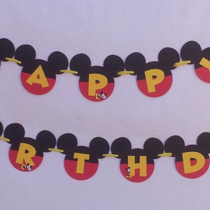 Pu&ograve; includere: Uno striscione di compleanno a due righe con sagome a forma di Mickey Mouse in nero, rosso e giallo. Le lettere HAPPY BIRTHDAY sono in giallo su cerchi rossi, affiancate da icone di teste di Mickey e pantaloncini rossi, su un nastro giallo.