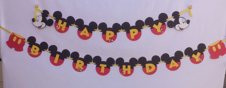 Pu&ograve; includere: Uno striscione di compleanno a due righe con sagome a forma di Mickey Mouse in nero, rosso e giallo. Le lettere HAPPY BIRTHDAY sono in giallo su cerchi rossi, affiancate da icone di teste di Mickey e pantaloncini rossi, su un nastro giallo.