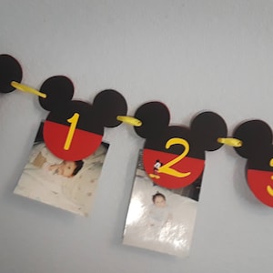 May include: A Mickey Mouse themed photo banner with red and black Mickey Mouse ears and yellow string. Each ear has a red and black circle with a number 1, 2, or 3 in yellow. The first ear has the letters "NB" in yellow. Each ear has a photo of a baby.