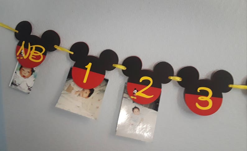 May include: A Mickey Mouse themed photo banner with red and black Mickey Mouse ears and yellow string. Each ear has a red and black circle with a number 1, 2, or 3 in yellow. The first ear has the letters "NB" in yellow. Each ear has a photo of a baby.