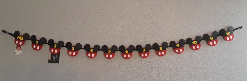 May include: A black and red Mickey Mouse themed banner with numbers 1 through 12 and "NB" for newborn. The banner is strung on a white wall.