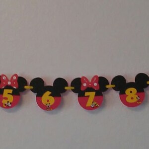 May include: A decorative banner featuring black mouse-ear silhouettes with red circular bases, each displaying a yellow number from NB to 12. Some sections include small square photographs. The pieces are connected by a yellow ribbon string.