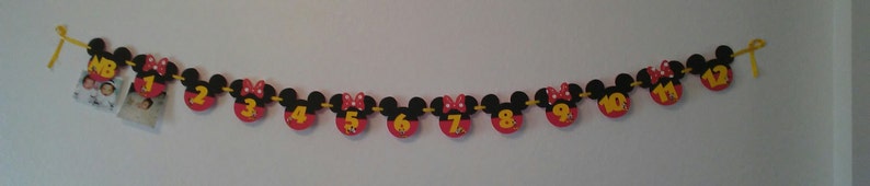 May include: A decorative banner featuring black mouse-ear silhouettes with red circular bases, each displaying a yellow number from NB to 12. Some sections include small square photographs. The pieces are connected by a yellow ribbon string.