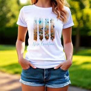May include: White t-shirt with a graphic design of colorful flowers and butterflies. The text "New Creation" is printed in blue on the front of the shirt.