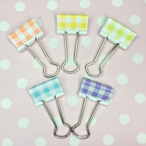 May include: Five binder clips with different coloured gingham patterns. The clips are arranged in a star shape on a pink background with white polka dots.