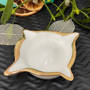Könnte beinhalten: Eine weiße Keramikschale mit goldenem Rand. Die Schale ist blumenförmig mit fünf Blütenblättern. Sie steht auf einer schwarzen Oberfläche.