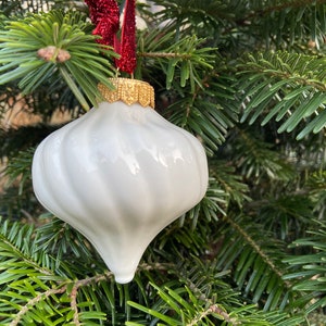 Op de afbeelding: Witte kerstbal van keramiek met een gouden rand en een swirl-ontwerp. De kerstbal hangt aan een rood lint aan een groene kerstboom.