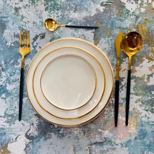 May include: A white ceramic dinner plate with a gold rim, set on a textured blue and white background. The plate is surrounded by gold and black flatware.